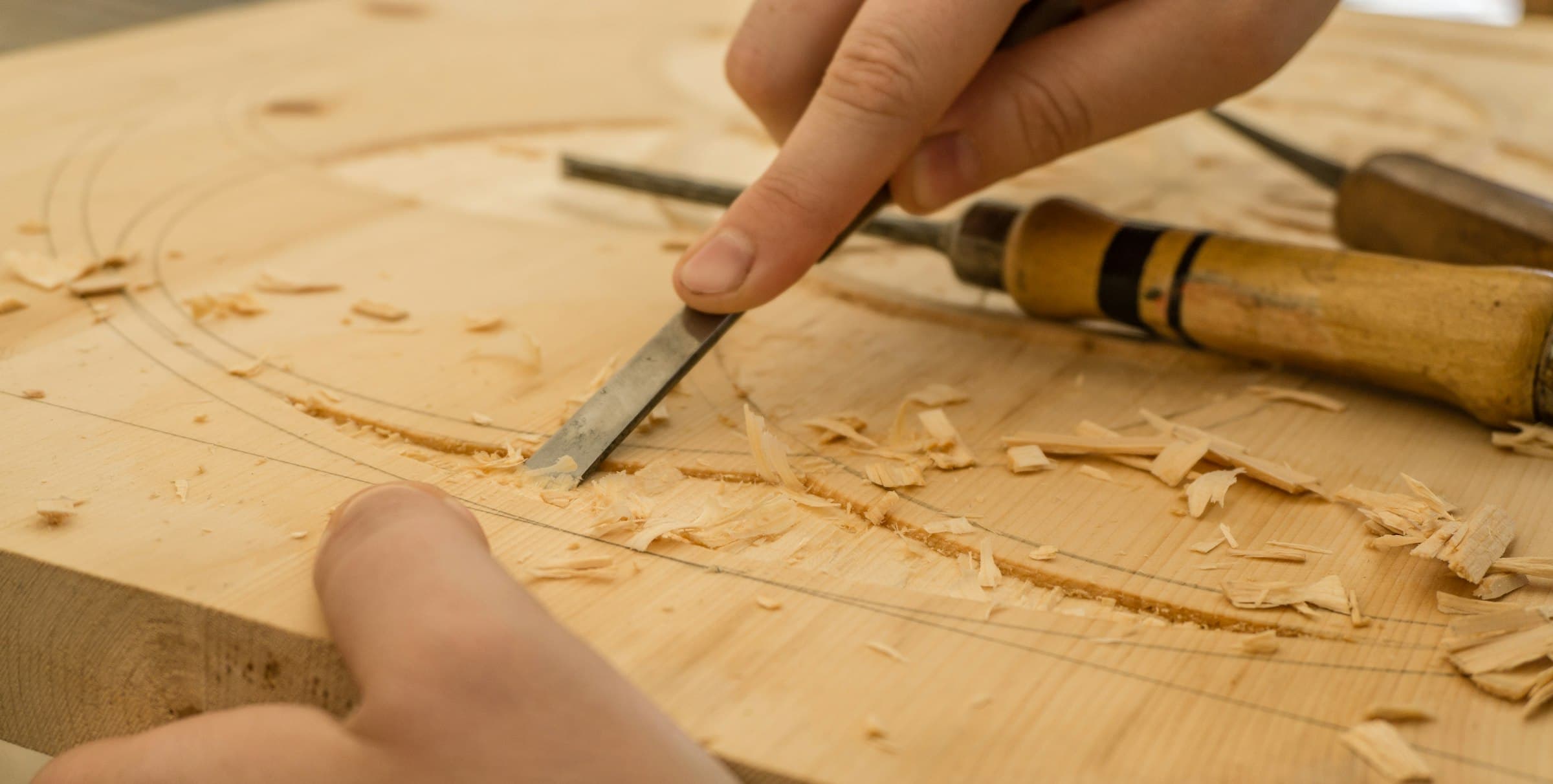 Hands guiding a chisel along a pencil line, shavings curled across the board