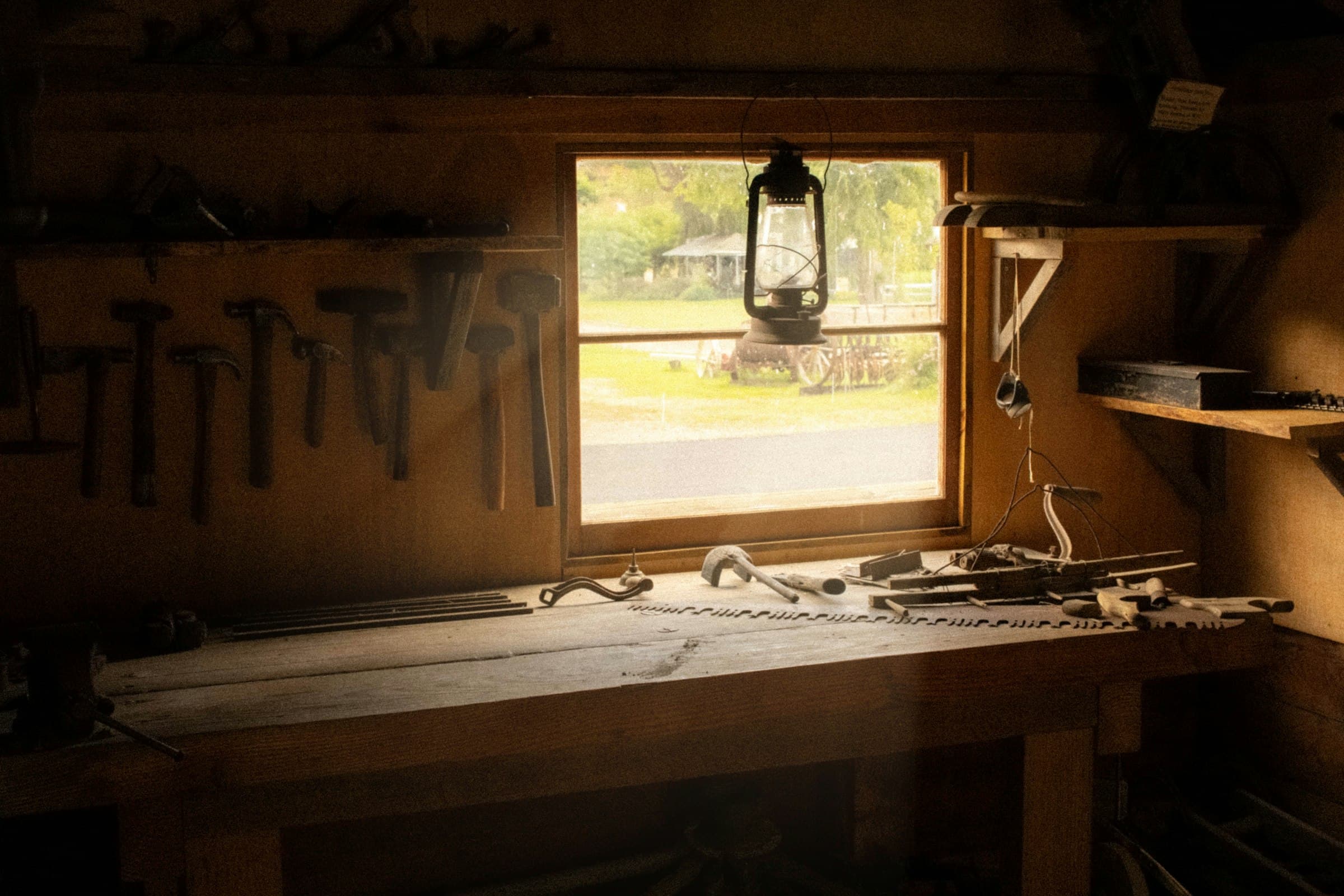 Sunlit workbench under a shop window — hammers and mallets hung on the wall, a kerosene lantern in the window, hand saws on the bench