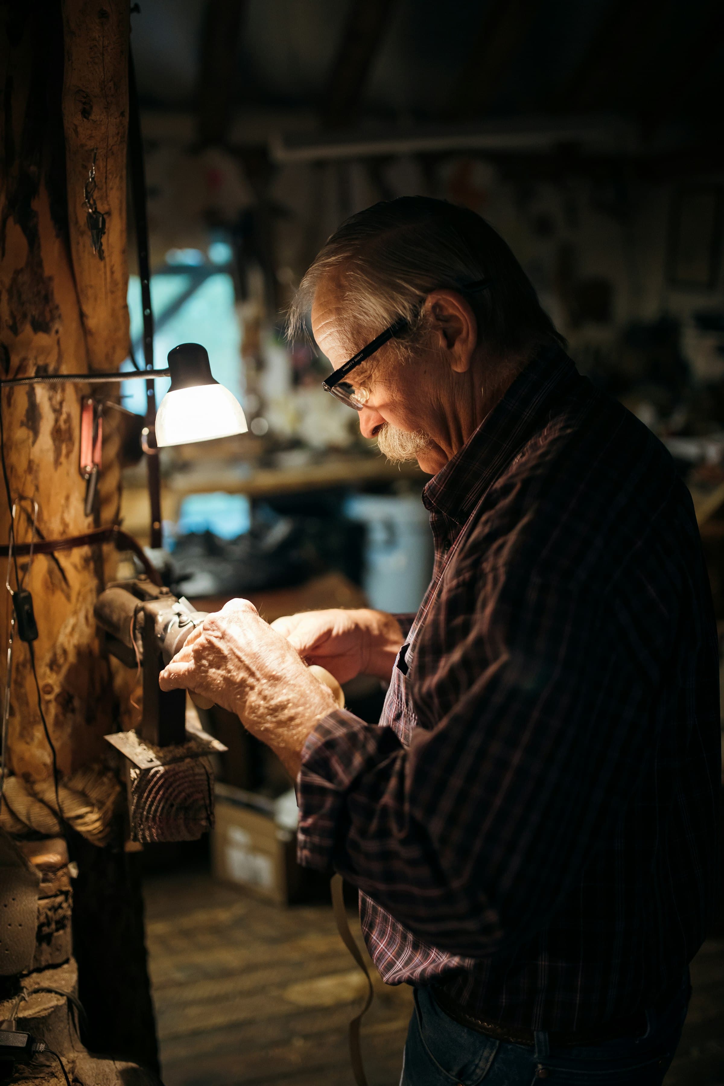 Ray Calloway at his bench — white hair, glasses, plaid shirt — absorbed in a repair under a work lamp